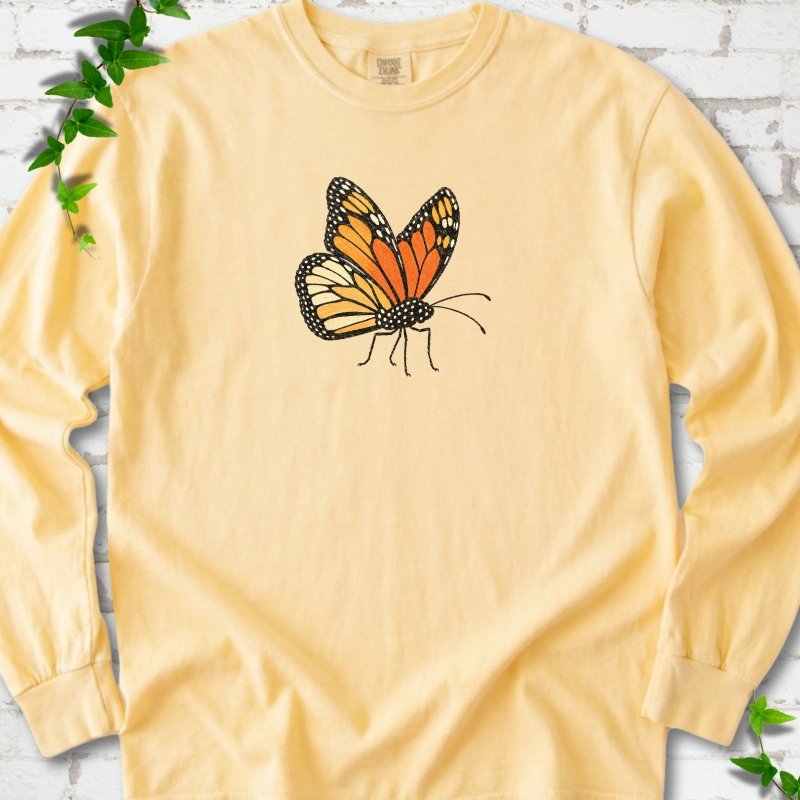 Butter yellow long sleeve T-shirt with one large orange and black monarch butterfly, wings raised, printed on the chest, flat lay on white brick with ivy