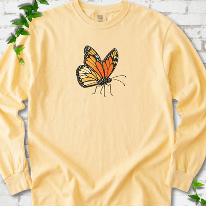 Butter yellow long sleeve T-shirt with one large orange and black monarch butterfly, wings raised, printed on the chest, flat lay on white brick with ivy