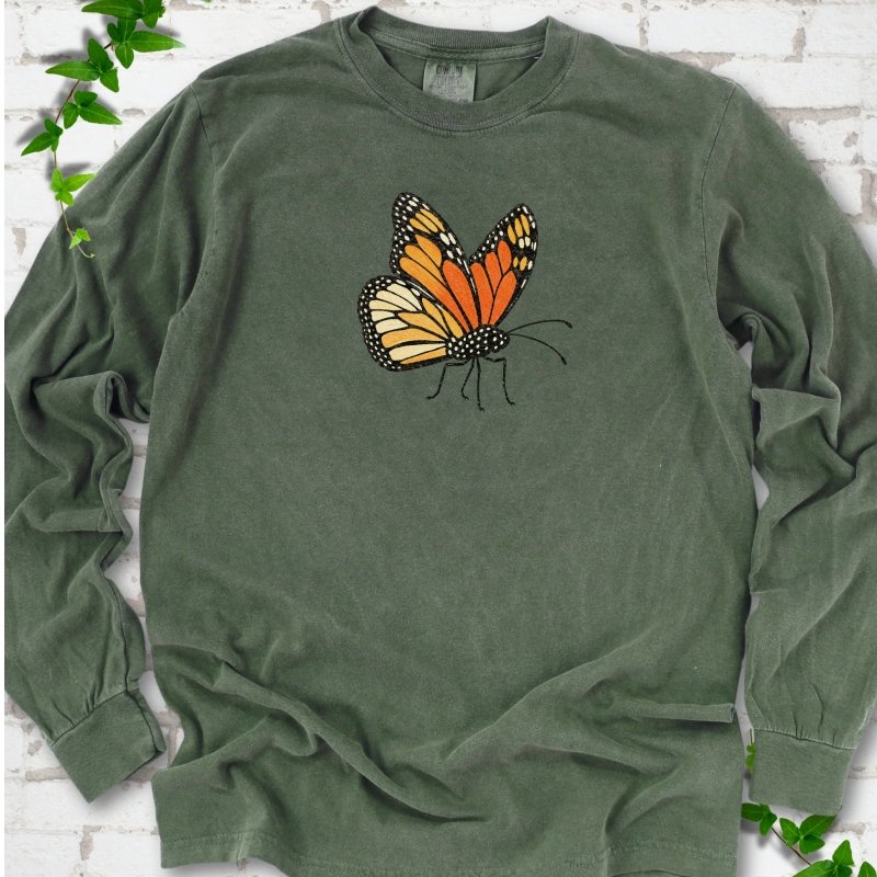 Hemp green long sleeve T-shirt with one large orange and black monarch butterfly, wings raised, printed on the chest, flat lay on white brick with ivy
