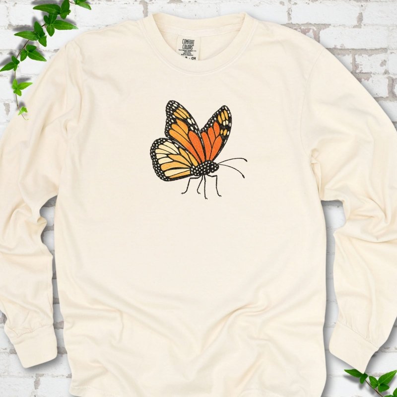 Ivory cream long sleeve T-shirt with one large orange and black monarch butterfly, wings raised, printed on the chest, flat lay on white brick with ivy