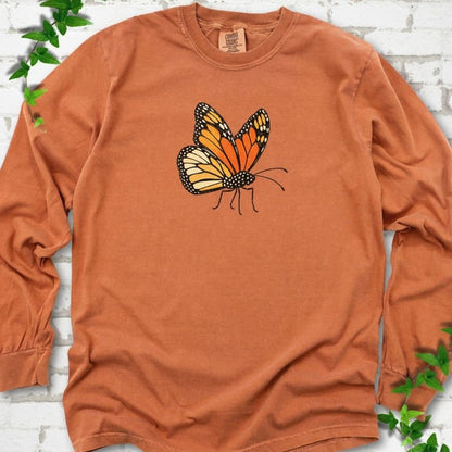 Yam burnt orange long sleeve T-shirt with one large orange and black monarch butterfly, wings raised, printed on the chest, flat lay on white brick with ivy
