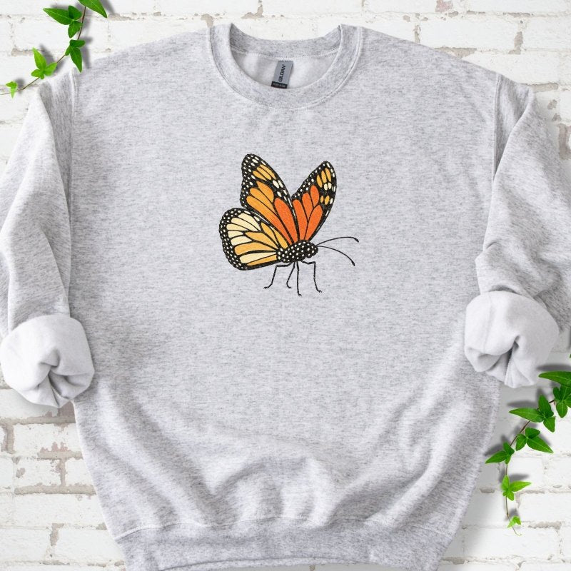 Ash light gray crewneck sweatshirt with one large orange and black monarch butterfly, wings raised, printed on the chest, flat lay on white brick with ivy