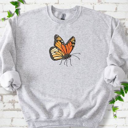 Ash light gray crewneck sweatshirt with one large orange and black monarch butterfly, wings raised, printed on the chest, flat lay on white brick with ivy