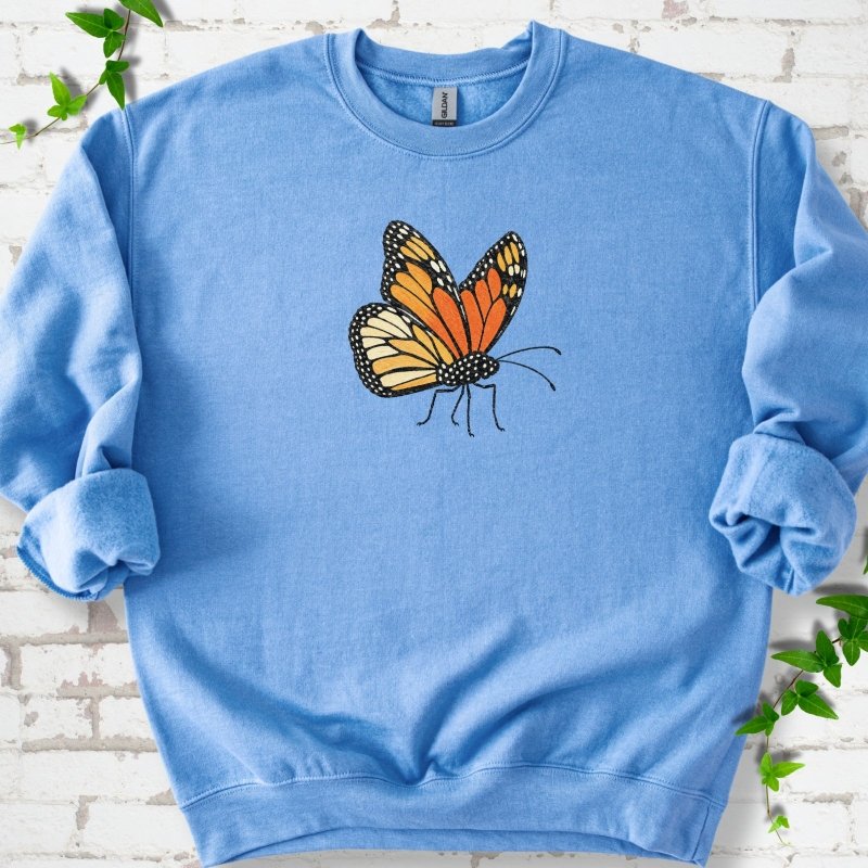 Carolina blue crewneck sweatshirt with one large orange and black monarch butterfly, wings raised, printed on the chest, flat lay on white brick with ivy