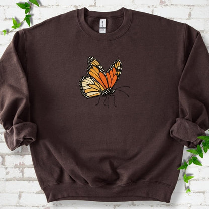 Dark chocolate brown crewneck sweatshirt with one large orange and black monarch butterfly, wings raised, printed on the chest, flat lay on white brick with ivy