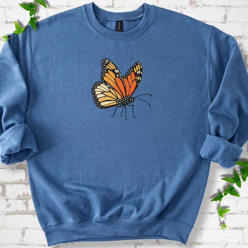 Indigo blue crewneck sweatshirt with one large orange and black monarch butterfly, wings raised, printed on the chest, flat lay on white brick with ivy