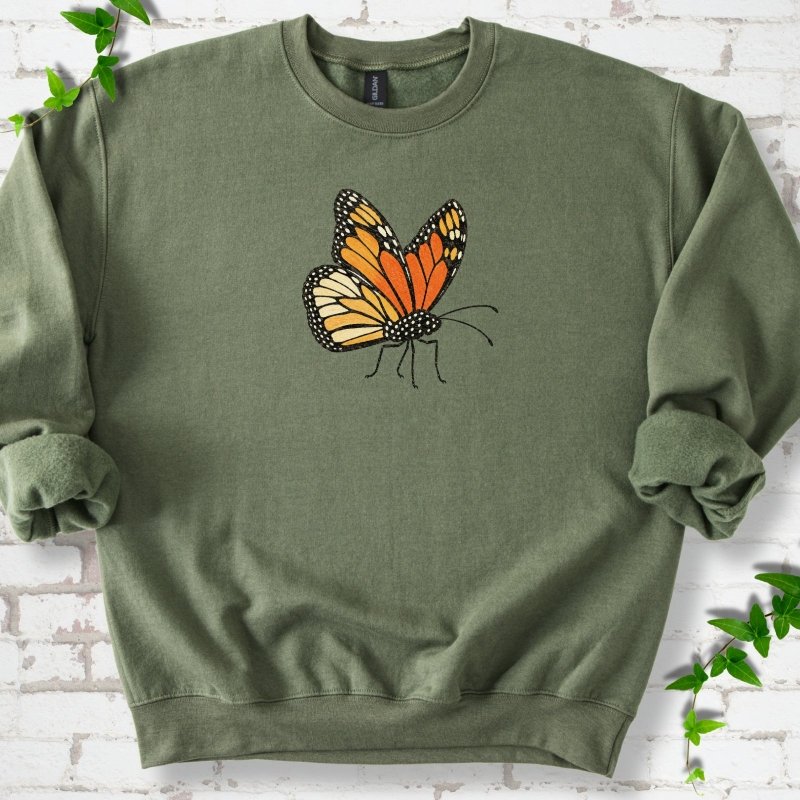 Military green crewneck sweatshirt with one large orange and black monarch butterfly, wings raised, printed on the chest, flat lay on white brick with ivy