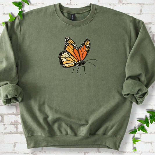 Military green crewneck sweatshirt with one large orange and black monarch butterfly, wings raised, printed on the chest, flat lay on white brick with ivy
