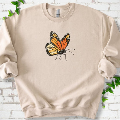 Sand beige crewneck sweatshirt with one large orange and black monarch butterfly, wings raised, printed on the chest, flat lay on white brick with ivy
