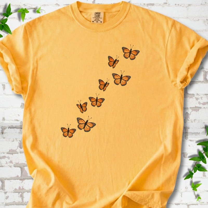 Citrus golden yellow short-sleeve T-shirt with eight orange monarch butterflies flying in a rising diagonal trail across the chest, flat lay on white brick with ivy
