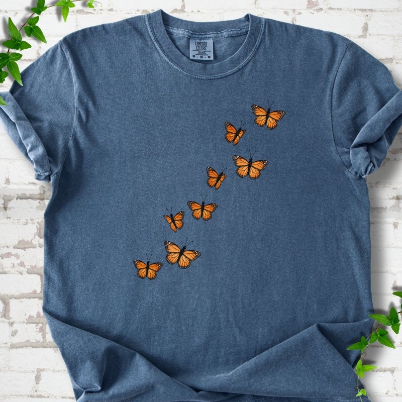 Denim blue short-sleeve T-shirt with eight orange monarch butterflies flying in a rising diagonal trail across the chest, flat lay on white brick with ivy