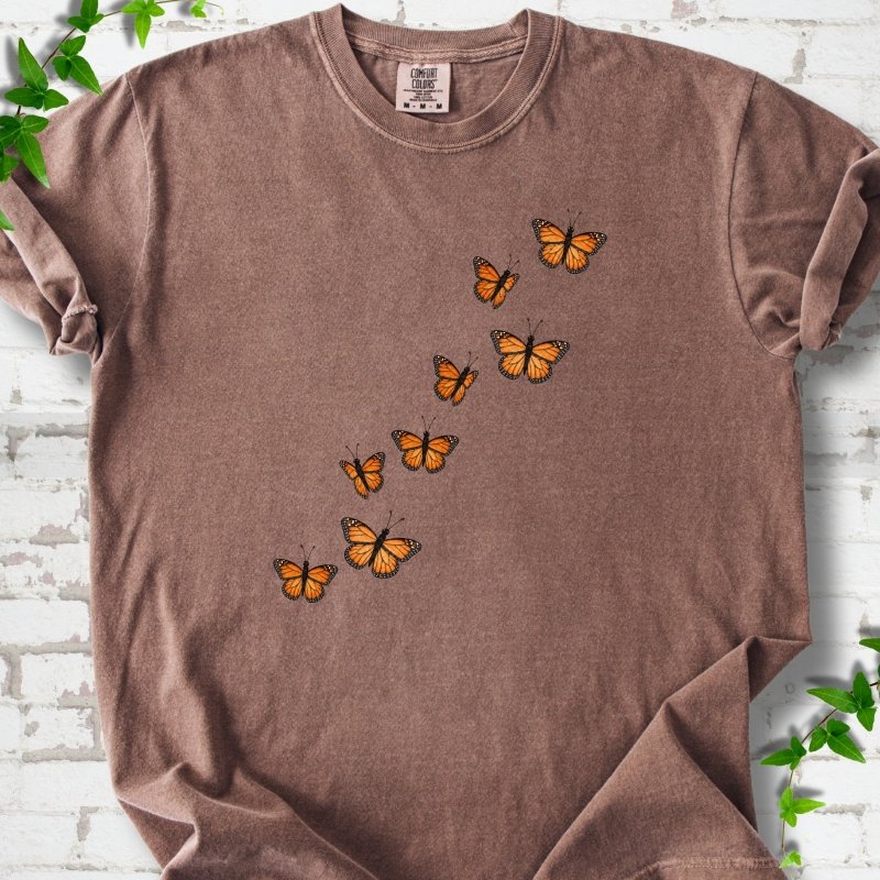 Espresso brown short-sleeve T-shirt with eight orange monarch butterflies flying in a rising diagonal trail across the chest, flat lay on white brick with ivy