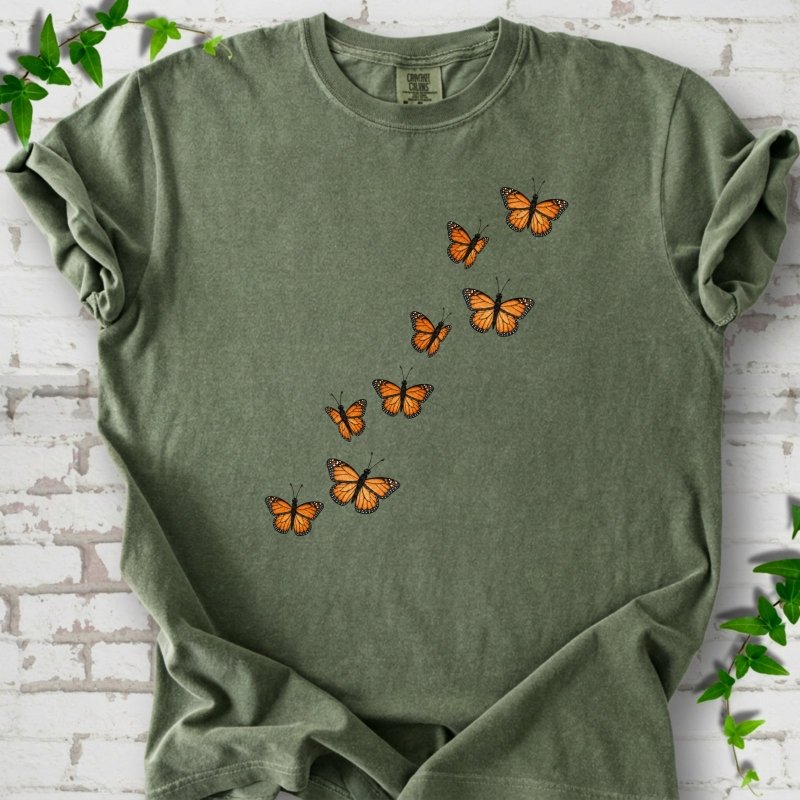 Moss green short-sleeve T-shirt with eight orange monarch butterflies flying in a rising diagonal trail across the chest, flat lay on white brick with ivy