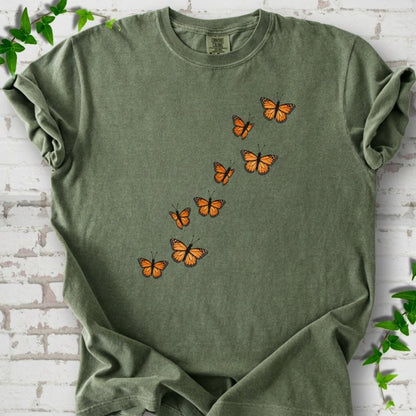 Moss green short-sleeve T-shirt with eight orange monarch butterflies flying in a rising diagonal trail across the chest, flat lay on white brick with ivy
