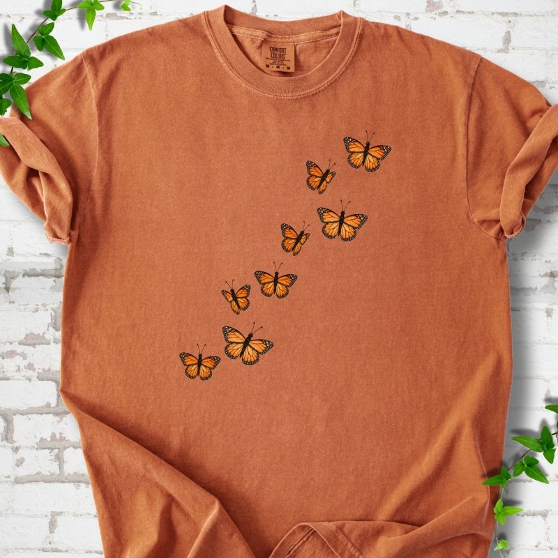 Yam burnt orange short-sleeve T-shirt with eight orange monarch butterflies flying in a rising diagonal trail across the chest, flat lay on white brick with ivy