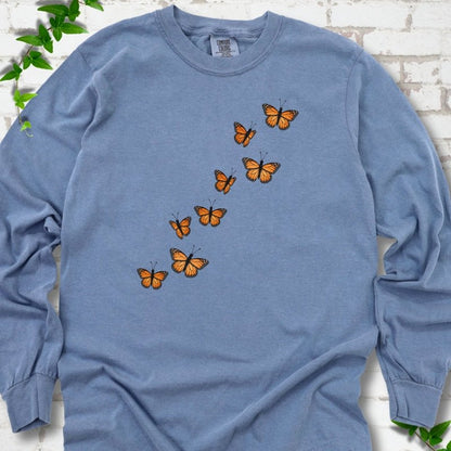 Blue Jean light blue long sleeve T-shirt with eight orange monarch butterflies flying in a rising diagonal trail across the chest, flat lay on white brick with ivy