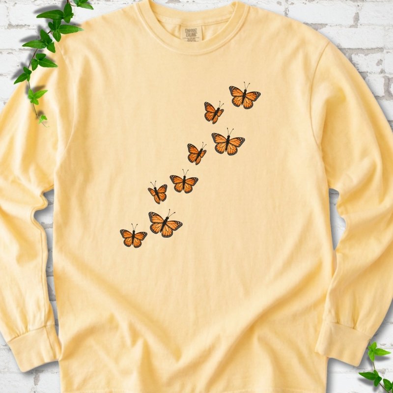 Butter yellow long sleeve T-shirt with eight orange monarch butterflies flying in a rising diagonal trail across the chest, flat lay on white brick with ivy
