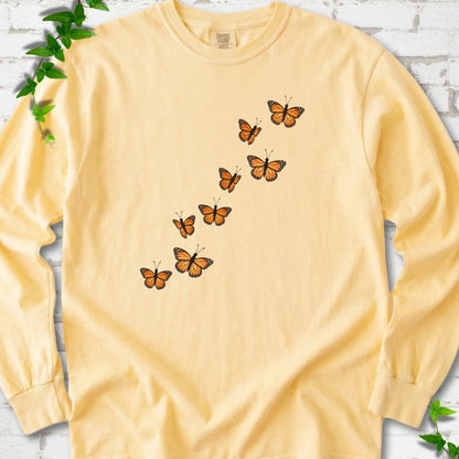 Butter yellow long sleeve T-shirt with eight orange monarch butterflies flying in a rising diagonal trail across the chest, flat lay on white brick with ivy