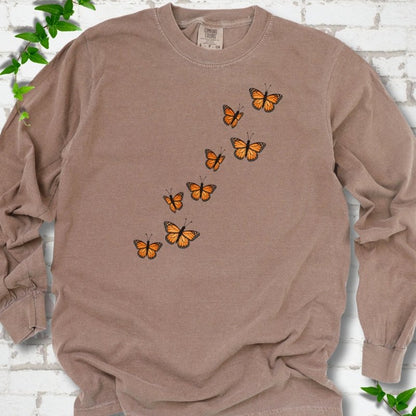 Espresso brown long sleeve T-shirt with eight orange monarch butterflies flying in a rising diagonal trail across the chest, flat lay on white brick with ivy