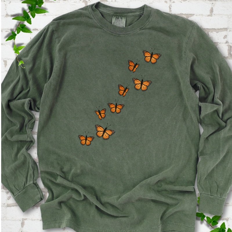 Hemp green long sleeve T-shirt with eight orange monarch butterflies flying in a rising diagonal trail across the chest, flat lay on white brick with ivy