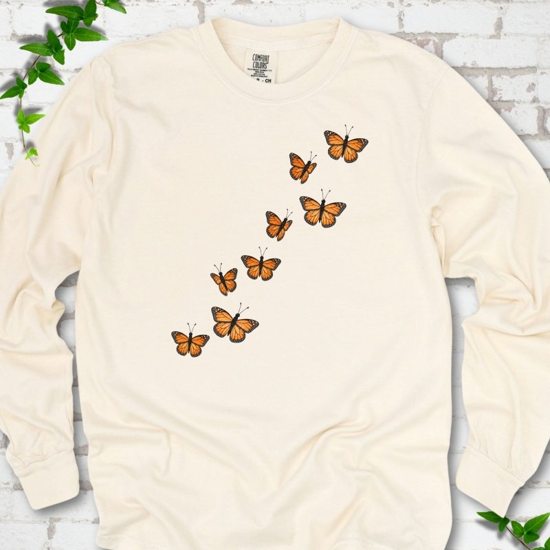 Ivory cream long sleeve T-shirt with eight orange monarch butterflies flying in a rising diagonal trail across the chest, flat lay on white brick with ivy