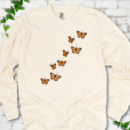 Ivory cream long sleeve T-shirt with eight orange monarch butterflies flying in a rising diagonal trail across the chest, flat lay on white brick with ivy
