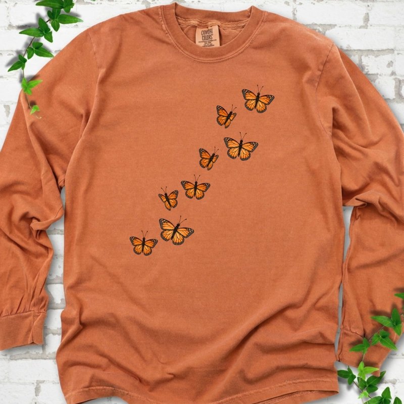 Yam burnt orange long sleeve T-shirt with eight orange monarch butterflies flying in a rising diagonal trail across the chest, flat lay on white brick with ivy