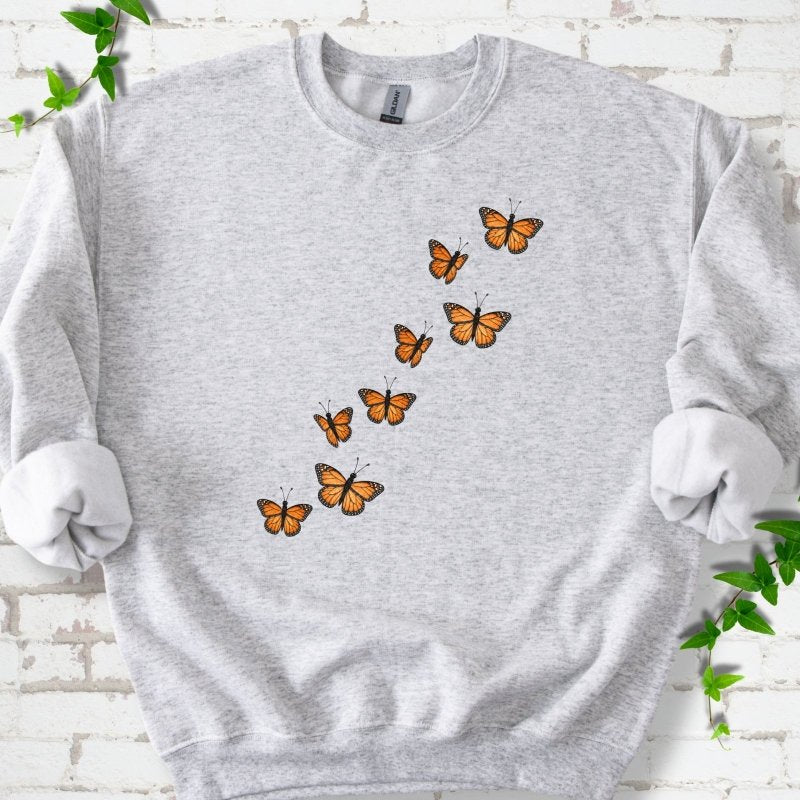 Ash light gray crewneck sweatshirt with eight orange monarch butterflies flying in a rising diagonal trail across the chest, flat lay on white brick with ivy
