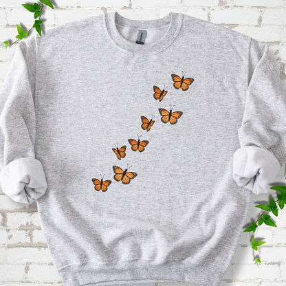 Ash light gray crewneck sweatshirt with eight orange monarch butterflies flying in a rising diagonal trail across the chest, flat lay on white brick with ivy