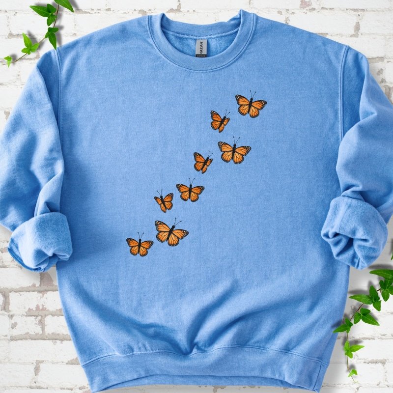 Carolina blue crewneck sweatshirt with eight orange monarch butterflies flying in a rising diagonal trail across the chest, flat lay on white brick with ivy