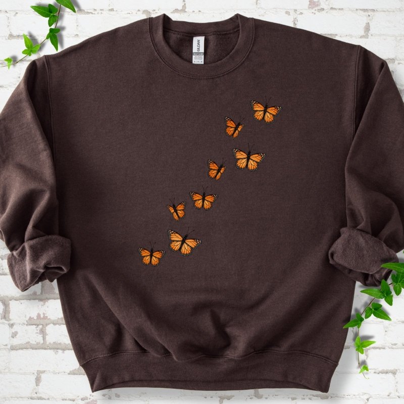 Dark chocolate brown crewneck sweatshirt with eight orange monarch butterflies flying in a rising diagonal trail across the chest, flat lay on white brick with ivy