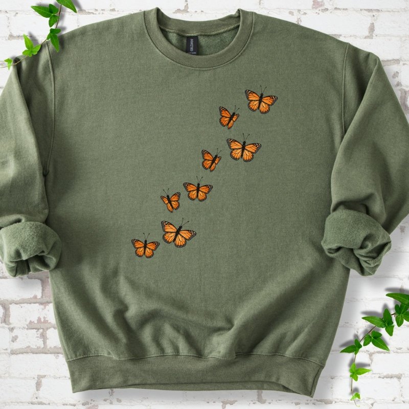 Military green crewneck sweatshirt with eight orange monarch butterflies flying in a rising diagonal trail across the chest, flat lay on white brick with ivy
