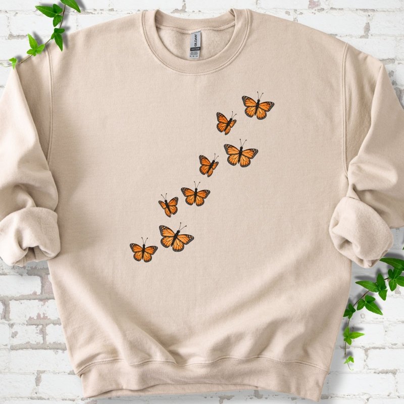 Sand beige crewneck sweatshirt with eight orange monarch butterflies flying in a rising diagonal trail across the chest, flat lay on white brick with ivy