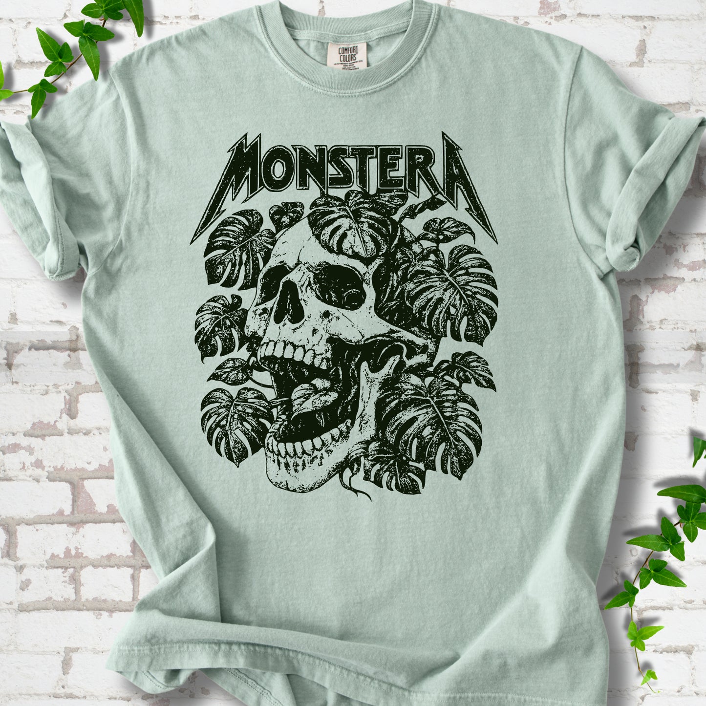 Bay Comfort Colors® T-shirt with distressed skull and monstera leaves beneath MONSTERA lettering, flat lay on white brick.