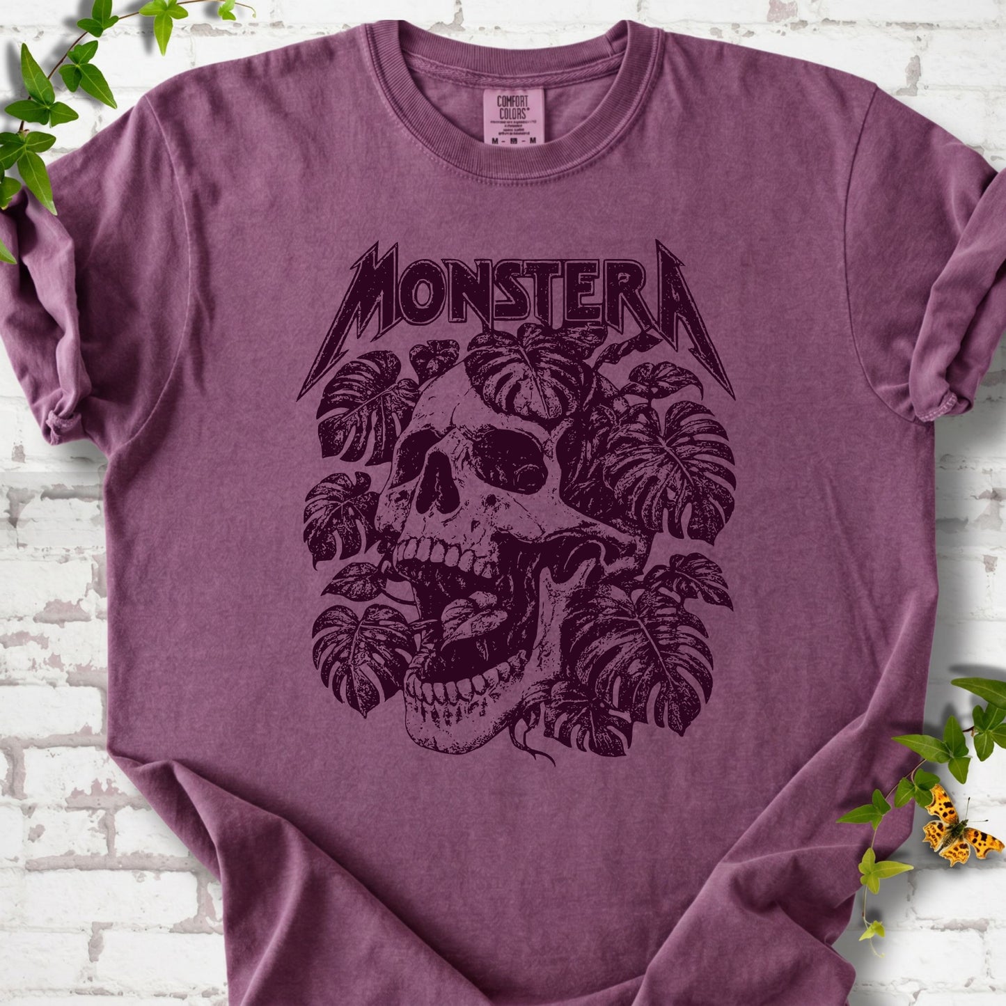 Berry Comfort Colors® T-shirt with distressed skull and monstera leaves beneath MONSTERA lettering, flat lay on white brick.
