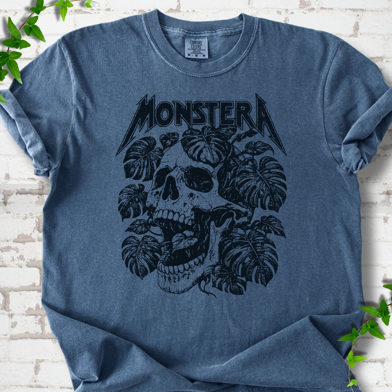 Denim Comfort Colors® T-shirt with distressed skull and monstera leaves beneath MONSTERA lettering, flat lay on white brick.