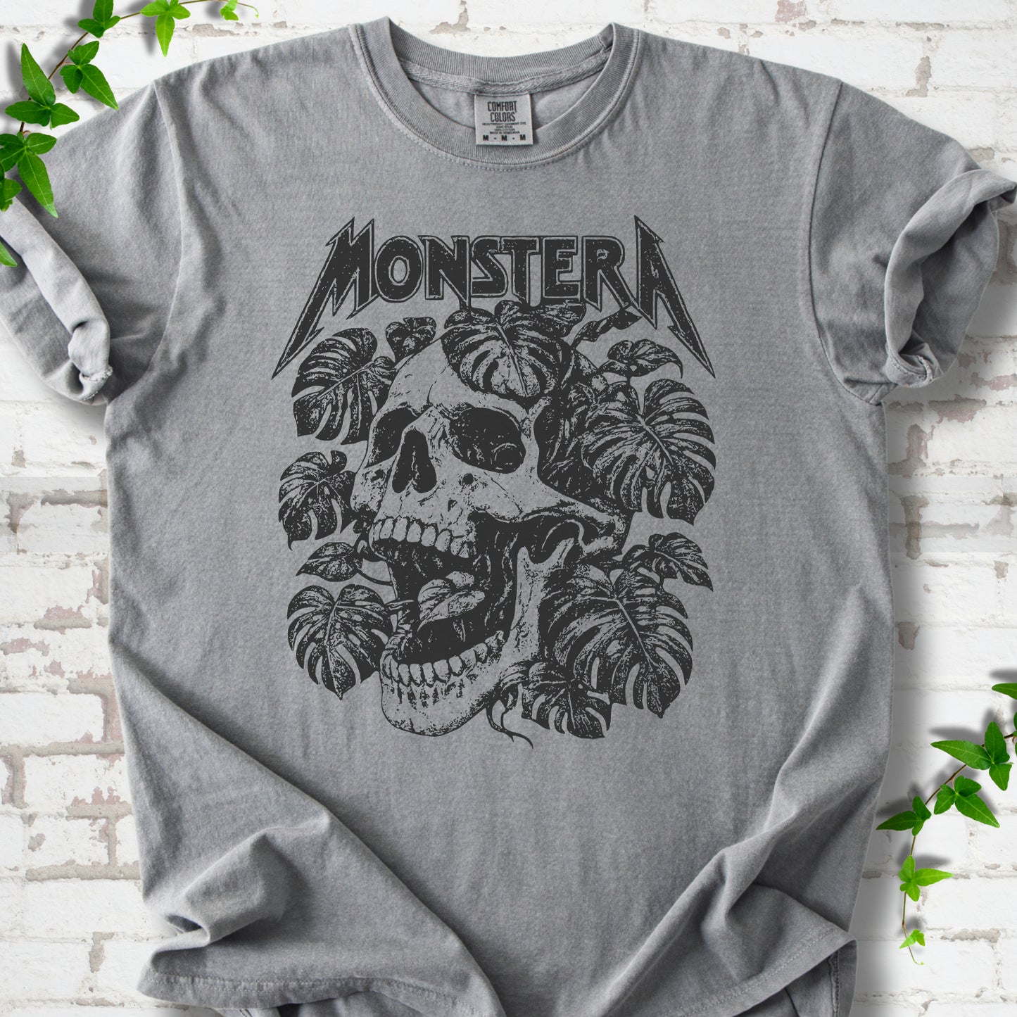 Grey Comfort Colors® T-shirt with distressed skull and monstera leaves beneath MONSTERA lettering, flat lay on white brick.