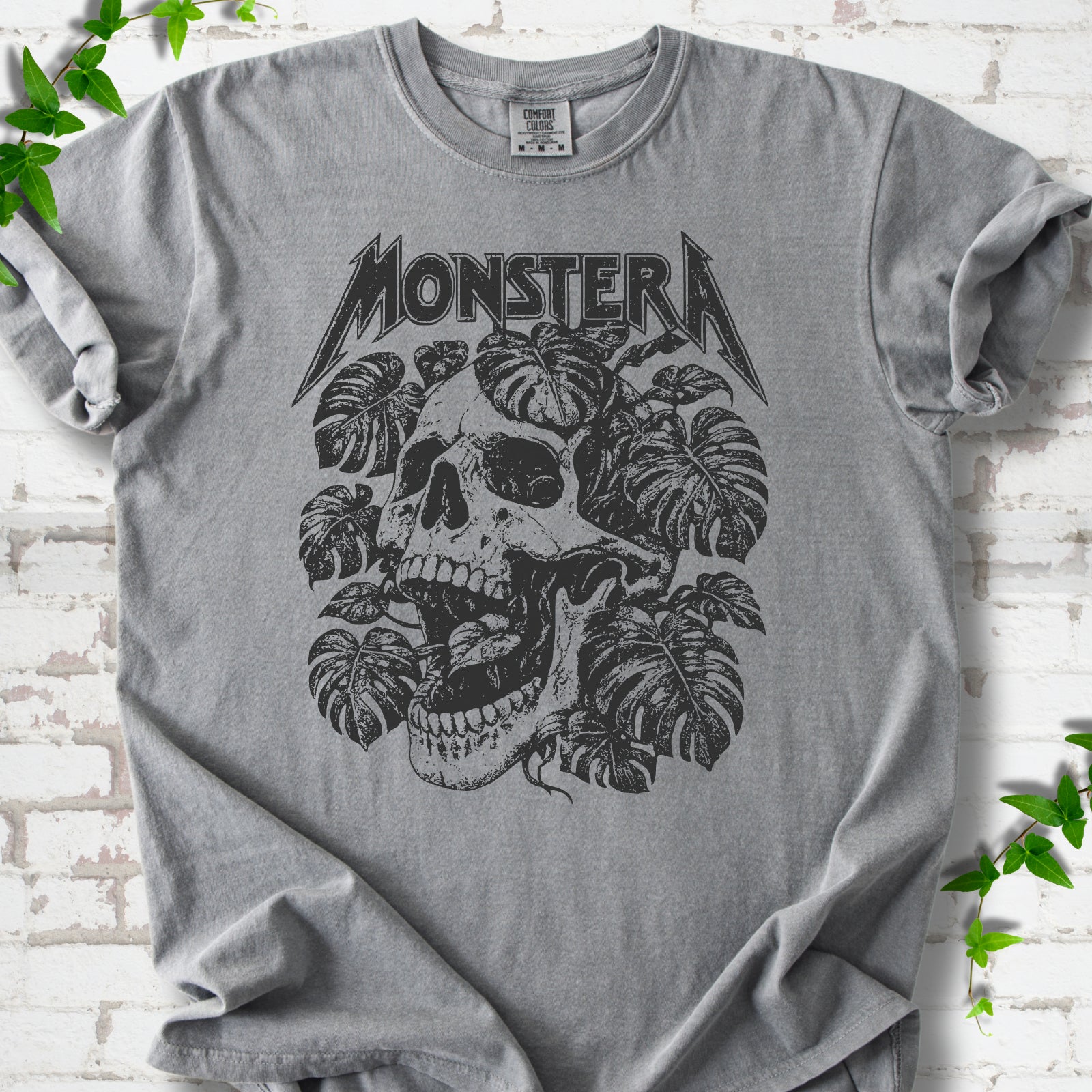 Grey Comfort Colors® T-shirt with distressed skull and monstera leaves beneath MONSTERA lettering, flat lay on white brick.