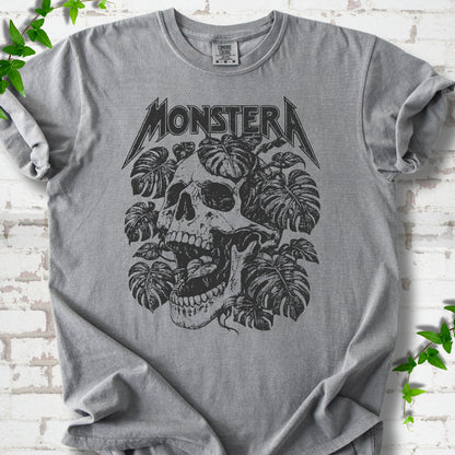 Grey Comfort Colors® T-shirt with distressed skull and monstera leaves beneath MONSTERA lettering, flat lay on white brick.