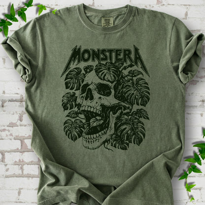 Moss Comfort Colors® T-shirt with distressed skull and monstera leaves beneath MONSTERA lettering, flat lay on white brick.