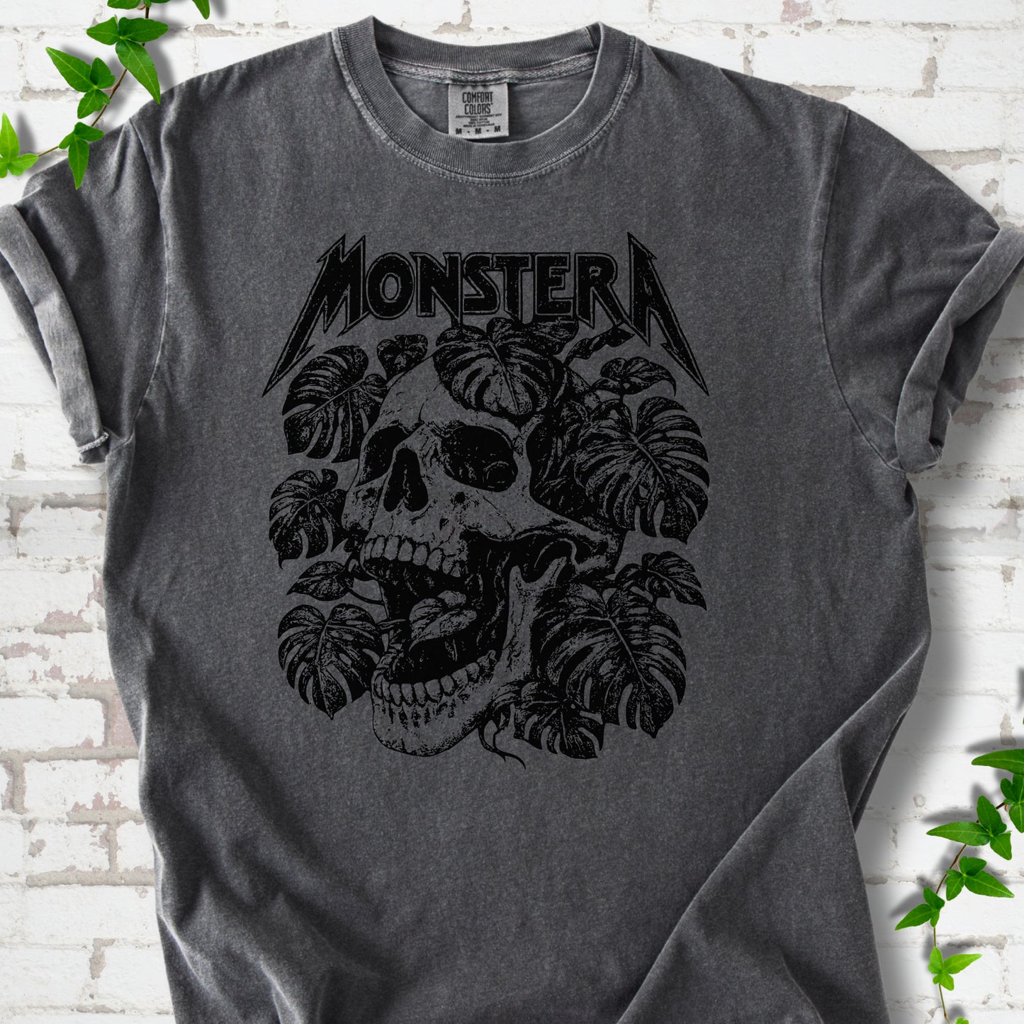 Pepper Comfort Colors® T-shirt with distressed skull and monstera leaves beneath MONSTERA lettering, flat lay on white brick.