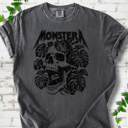 Pepper Comfort Colors® T-shirt with distressed skull and monstera leaves beneath MONSTERA lettering, flat lay on white brick.
