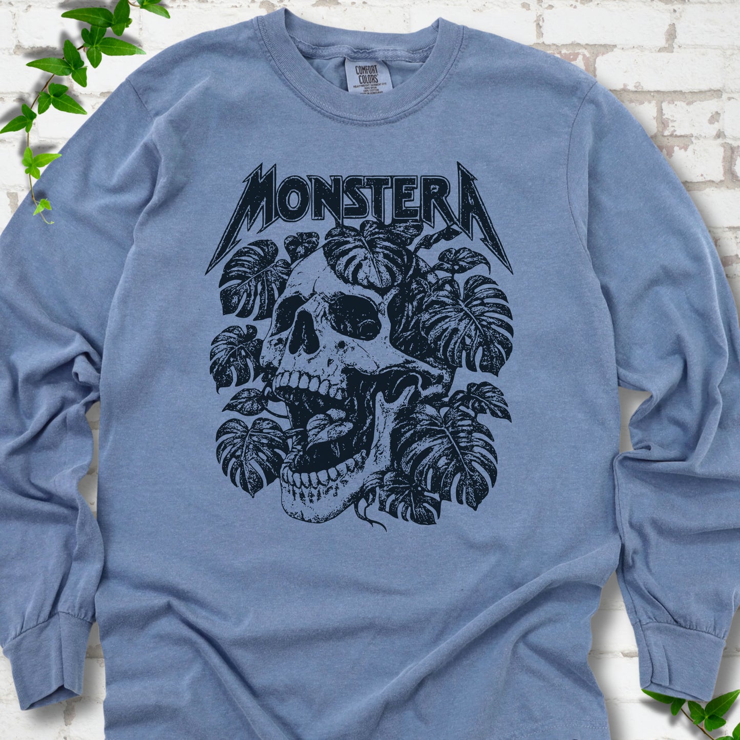 Blue Jean Comfort Colors® long sleeve T-shirt with distressed skull and monstera leaves beneath MONSTERA lettering, flat lay on white brick.