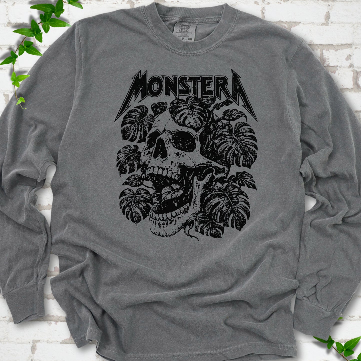 Pepper Comfort Colors® long sleeve T-shirt with distressed skull and monstera leaves beneath MONSTERA lettering, flat lay on white brick.