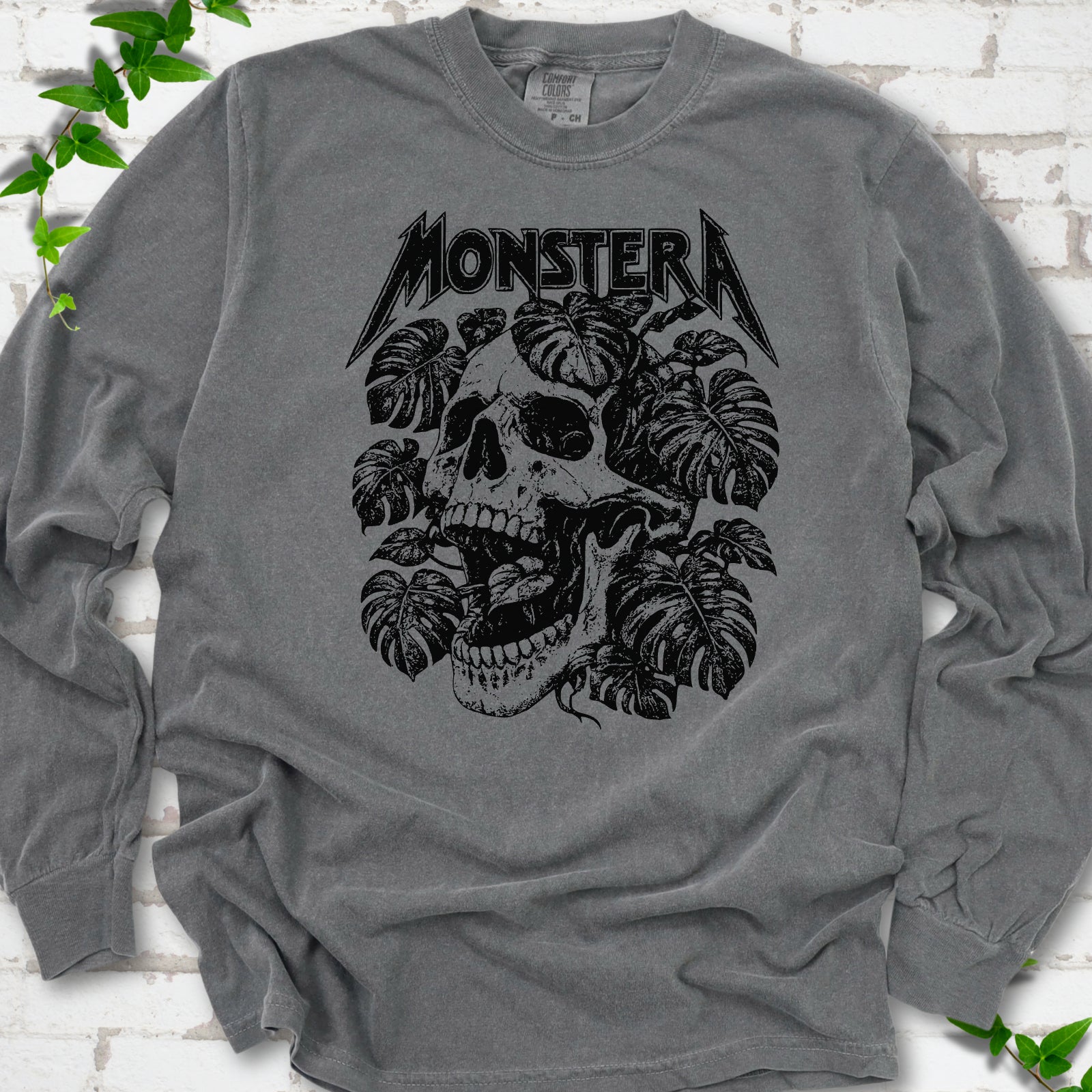 Pepper Comfort Colors® long sleeve T-shirt with distressed skull and monstera leaves beneath MONSTERA lettering, flat lay on white brick.