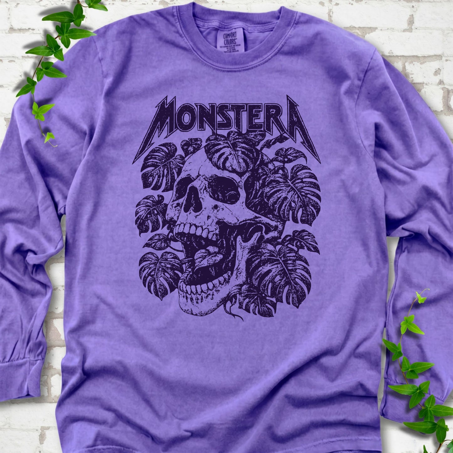 Violet Comfort Colors® long sleeve T-shirt with distressed skull and monstera leaves beneath MONSTERA lettering, flat lay on white brick.
