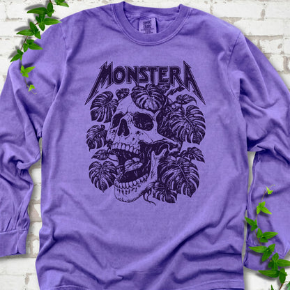 Violet Comfort Colors® long sleeve T-shirt with distressed skull and monstera leaves beneath MONSTERA lettering, flat lay on white brick.