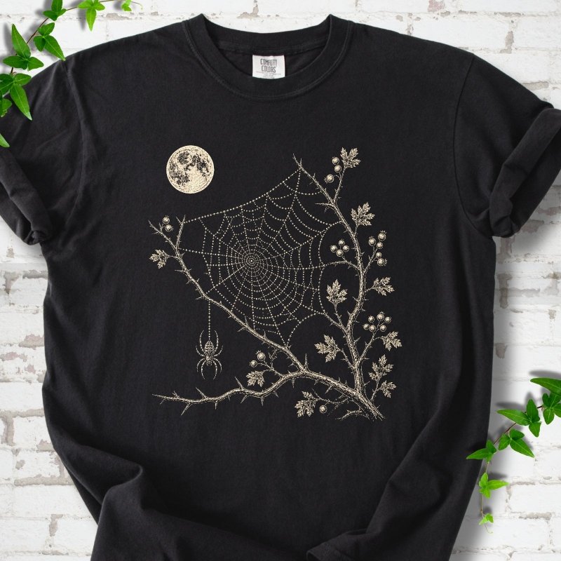 Black Comfort Colors® T-shirt flat lay featuring a cream moon, spider web, berry branches and hanging spider design.