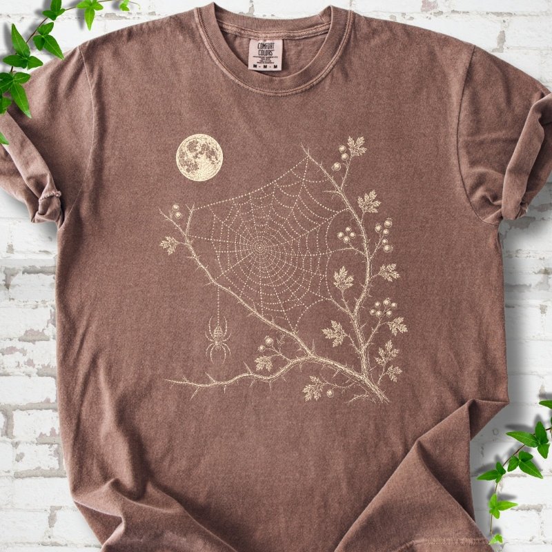 Espresso Comfort Colors® T-shirt flat lay featuring a cream moon, spider web, berry branches and hanging spider design.