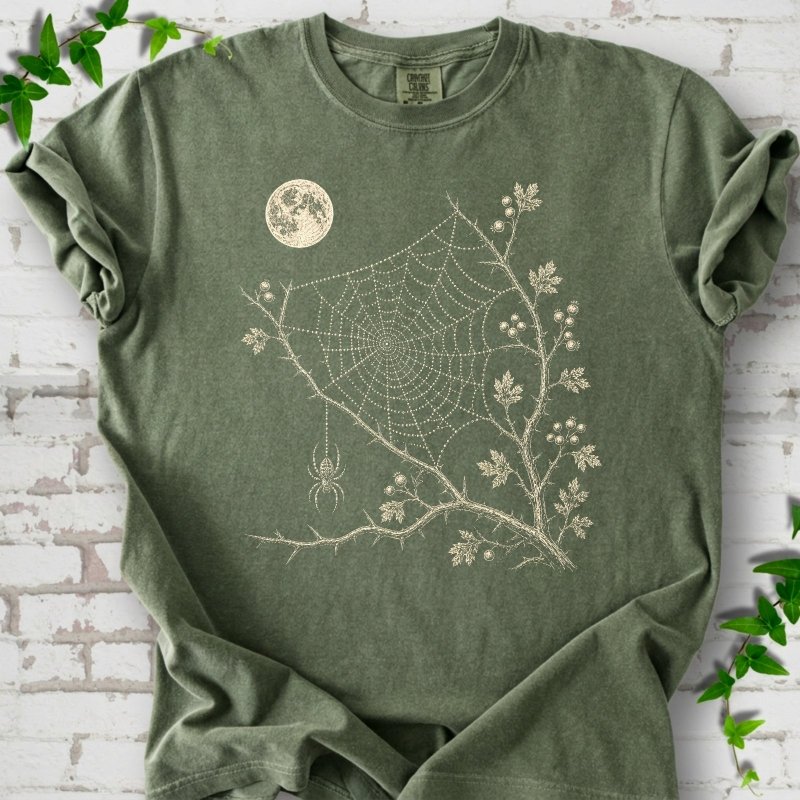 Moss Comfort Colors® T-shirt flat lay featuring a cream moon, spider web, berry branches and hanging spider design.